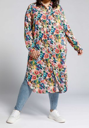 Personne portant une longue robe-chemise fleurie et colorée sur un jean bleu clair et des baskets blanches, debout, jambes écartées sur un fond uni.