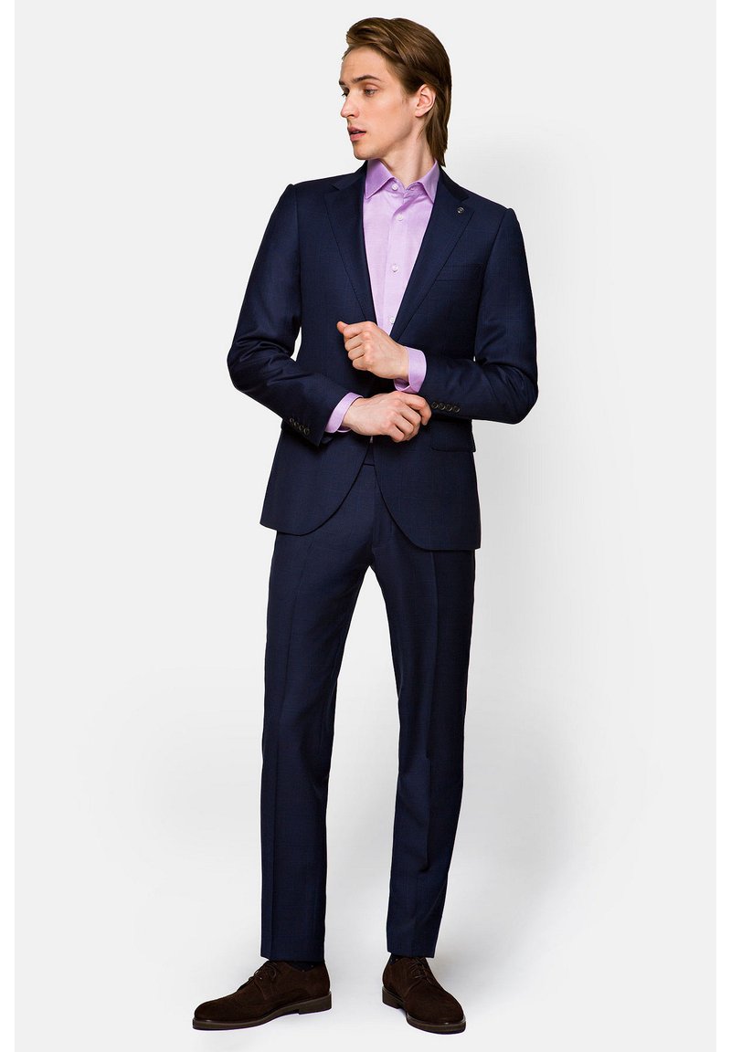 Granatowy garnitur o slim fit, stylizowany z jasnofioletową koszulą. Posiada subtelny wzór, jednorzędową marynarkę i ciemno-brązowe zamszowe buty.