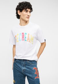 Λευκό βαμβακερό μπλουζάκι με πολύχρωμη επιγραφή "ICE CREAM", συνδυασμένο με μπλε τζιν παντελόνι που διαθέτει κόκκινη κεντητή λεπτομέρεια και εφαρμογές πατς.