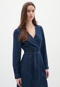 Jeanswrapklänning i mörkblått med långa ärmar, djup V-ringning och knut vid midjan. Synliga sömmar och sidofickor ger extra detalj.