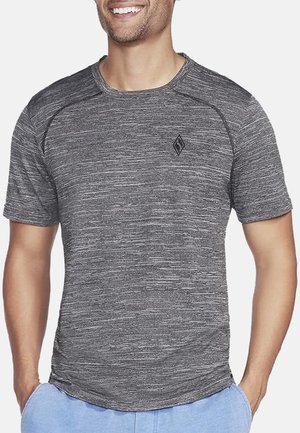 Mann trägt ein kurzärmliges, grau meliertes sportliches T-Shirt mit einem kleinen schwarzen Diamant-Logo auf der Brust, Hände in den Taschen einer hellblauen Hose.