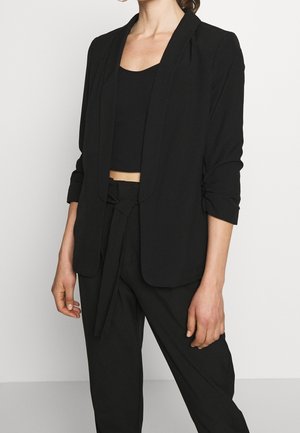 Mujer vistiendo un top negro corto, blazer negro con las mangas remangadas y pantalones negros de tiro alto con cinturón atado, de pie frente a un fondo liso.