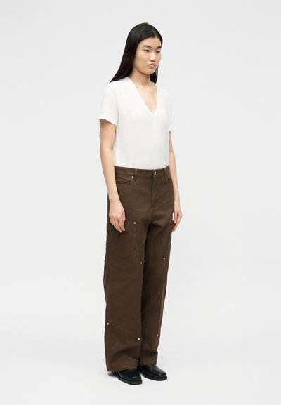 Γυναίκα που φοράει λευκό τοπ με κοντά μανίκια και V-λαιμόκοψη, καφέ παντελόνι με φαρδιά μπατζάκια και διακοσμητικά πριτσίνια, καθώς και μαύρα παπούτσια, στέκεται μπροστά σε ένα απλό φωτεινό φόντο.