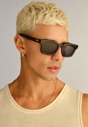 Młody mężczyzna z krótkimi, wybielonymi włosami, noszący okulary przeciwsłoneczne w oprawkach w panterkę, złotą kolczyk w kształcie koła, złoty łańcuszek i kremowy top bez rękawów.