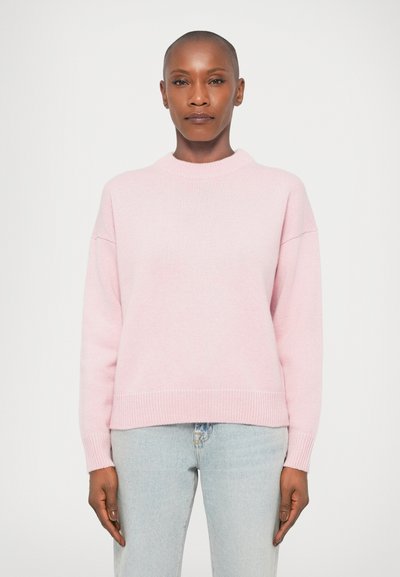 Femme avec les cheveux courts portant un pull pastel rose et un jean bleu clair, se tenant devant un fond blanc uni.