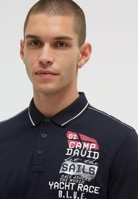 Marineblaues Poloshirt mit Kragen, weißen Akzenten und besticktem Text mit Zahlen und Phrasen, die sich auf Segeln und Yacht-Rennen beziehen.