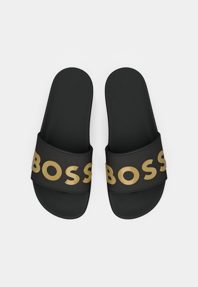 Claquettes noires avec une large lanière présentant des lettres dorées en relief épelant "BOSS". Texture lisse avec des bords arrondis.