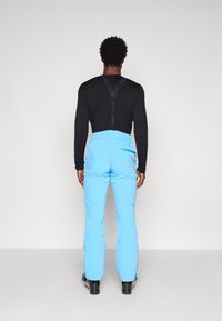 Šviesiai mėlynos slidinėjimo kelnės su reguliuojamomis petnešomis, užtrauktuku užpakalinėje kišenėje ir tiesiu šlaunų siluetu, derinamos su juoda ilgomis rankovėmis marškiniais.