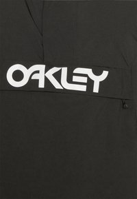 Czarna kurtka z dużym białym logo "OAKLEY" na piersi, o gładkiej fakturze materiału i sznurku ściągającym u dołu.