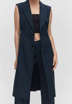 Ærmeløs navy lang blazer med to knapper båret over sort cropped top og matchende navy bukser på en stående person.