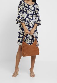 Brun flätad läderhandväska med en böjd topp, hålls av en person klädd i en blommönstrad klänning med vita och gula blommor på en marinblå bakgrund.