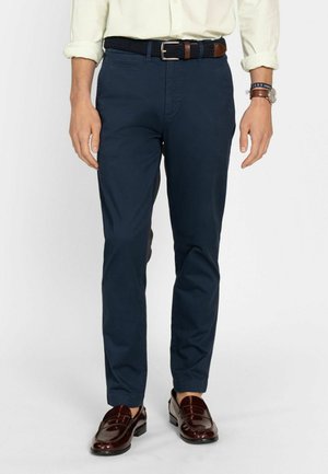 Hombre con pantalones chinos azul marino, cinturón trenzado oscuro, camisa de color claro arremangada, mocasines de cuero marrón y reloj de pulsera con pulsera.