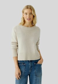 Beige gestrickter Pullover mit einem gerippten Saum und Ärmeln, ausgestattet mit einer Kängurutasche und einem runden Ausschnitt, kombiniert mit blauen Jeans mit weitem Bein.
