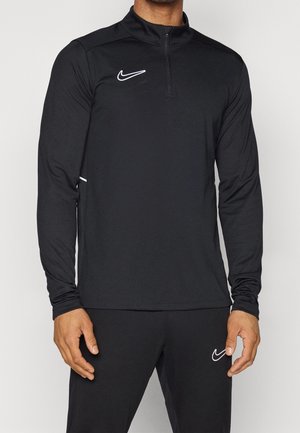 Mężczyzna noszący czarną sportową bluzę Nike z długim rękawem i suwakiem do ćwiartki oraz dopasowane czarne spodnie, obie z białymi logo Nike swoosh.
