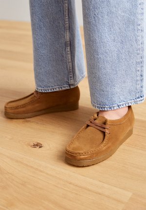 Brown suede shoes with a rounded toe and tan rubber sole, featuring a simple lace design, paired with light blue denim pants.