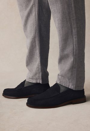 Gray textured pants worn with dark navy suede loafers and dark gray socks on a neutral light background.
