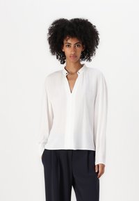 Camicia bianca con scollo a V e dettagli con bottoni, caratterizzata da maniche lunghe e una consistenza leggera e liscia. Abbinata a pantaloni scuri e larghi.