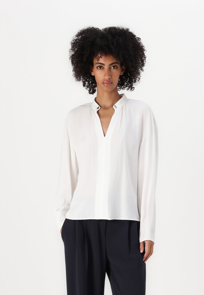Camicia bianca con scollo a V e dettagli con bottoni, caratterizzata da maniche lunghe e una consistenza leggera e liscia. Abbinata a pantaloni scuri e larghi.