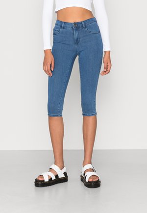 Woman wearing blue cropped skinny jeans, white long-sleeve cropped top, and white sandals with black thick soles standing on gray floor.