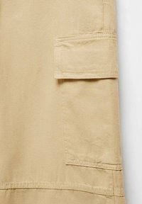 Nærbillede af beige stof med en klaplomme med syede kanter, tekstureret overflade og sømme. Klassisk cargo-design.