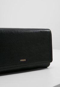 Portafoglio in pelle nera con finitura testurizzata, dotato del marchio dorato "FOSSIL", chiusura a scatto e dettagli di cucitura a contrasto.