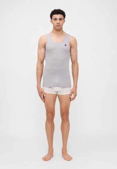 Débardeur à côtes gris clair avec de larges bretelles et un petit logo foncé, associé à des briefs blancs, modèle se tenant pieds nus contre un fond neutre.