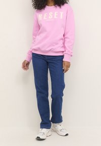 Persona vestida con un sudadera rosa con el texto "RESET", jeans azules y zapatillas blancas, de pie contra un fondo liso claro.