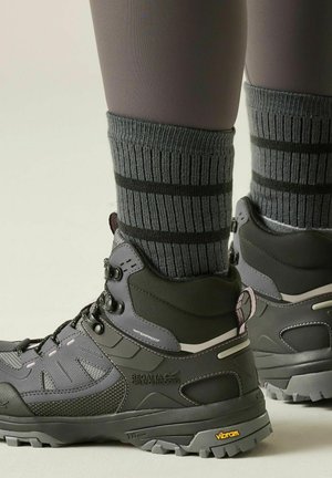 Nogi w szarych skarpetkach w paski i czarnych wodoodpornych butach trekkingowych z podeszwami Vibram na jasnym tle.