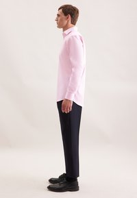 Uomo in piedi di lato, indossa una camicia a maniche lunghe rosa chiaro, pantaloni scuri e scarpe nere, su uno sfondo semplice e chiaro.