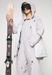 Îmbrăcăminte de schi care include o jachetă izolată gri deschis și un costum integrat asortat, cu ochelari de schi negri și o schi de culoare maro cu fixări negre.