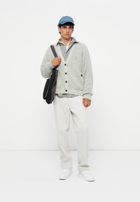 Gray knit cardigan with black buttons, layered over a striped shirt. White trousers and sneakers complete the outfit, with a blue cap on the head.