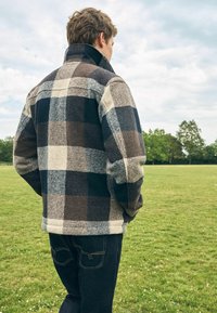 Man wearing a blue, brown, and cream checkered jacket and dark jeans standing in a grassy field with trees and cloudy sky in the background.