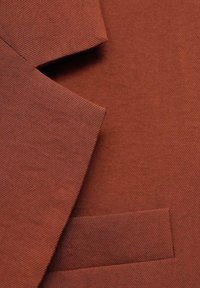 Tela color óxido con solapa de muesca y un bolsillo en el pecho, mostrando una fina textura de tejido diagonal en una prenda a medida.