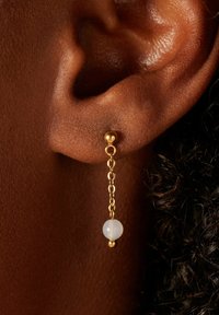 Boucles d'oreilles en chaîne dorée présentant une perle blanche, suspendue juste en dessous du lobe de l'oreille. Le design est délicat avec des surfaces lisses et réfléchissantes.