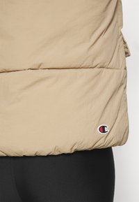 Doudoune beige avec une texture légèrement froissée. Présente un patch logo avec des accents rouges et noirs à l'ourlet. Legging noir en dessous.