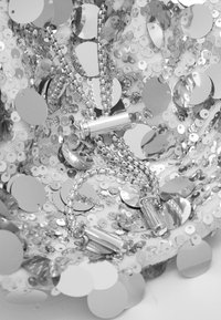 Srebrna tkanina z cekinami ma okrągłe, błyszczące cekiny o odbijającej powierzchni. Srebrne łańcuchy i metaliczne detale splatają się w projekcie.