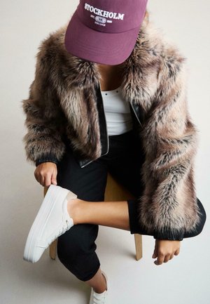 Kabát z umelej kožušiny v hnedorýhlym a sivých tónoch s čiernymi koženými akcentmi, doplnený o bielu čiapku a platformové tenisky. Drevená stolička v pozadí.