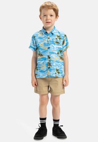 Korte mouwen, blauw patroonhemd met palmbomen en tekst, gecombineerd met beige shorts en zwarte sneakers met witte strepen.
