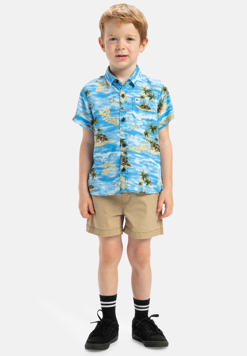 Korte mouwen, blauw patroonhemd met palmbomen en tekst, gecombineerd met beige shorts en zwarte sneakers met witte strepen.