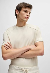 T-shirt sportivo color beige chiaro con maniche corte, tessuto testurizzato, scollatura a costine e dettagli sulle spalle; il modello posano con le braccia incrociate.