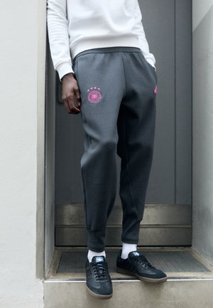 Pantalones jogger grises con un emblema rosa que presenta cuatro estrellas y un logo, cinturilla elástica, bolsillos laterales, combinados con zapatillas negras y calcetines blancos.