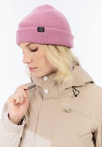 Rosa gestrickte Mütze mit umgeschlagenem Bündchen und schwarzem Etikett, kombiniert mit einer beigen Winterjacke mit Reißverschlusstasche und Kordelzügen.