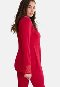 Rode blouse met lange mouwen en doorschijnende accenten bij de manchetten, een soepele textuur en een getailleerde pasvorm. Gedragen met bijpassende rode broek.