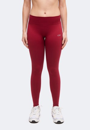 Femme portant des leggings rouges taille haute avec un petit logo blanc "ICANIWILL" sur la cuisse et des baskets blanches, debout devant un fond uni.