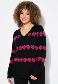 Donna con i capelli ricci che indossa un maglione nero a V con tre file di cuori rosa, in piedi davanti a uno sfondo bianco.