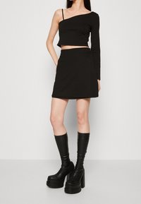 Femme portant un crop top noir à une épaule, une mini-jupe noire et de grandes bottes plateformes noires, debout devant un fond uni.