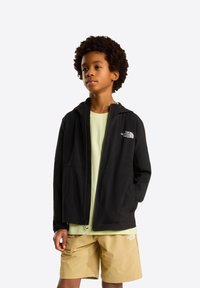 Jongen met krullend haar die een zwart zip-up North Face-jas draagt over een lichtgroen shirt en beige North Face-short, staand met één hand in zijn zak.