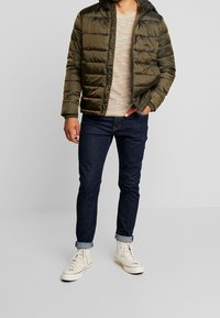 Giacca imbottita verde oliva con cappuccio e chiusura con zip; indossata sopra un maglione beige chiaro e jeans blu scuro; completata con sneakers bianche.