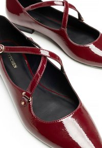 Scarpe basse in pelle laccata rossa con cinturini incrociati e piccoli accenti dorati. Punta rotonda, interno nero e tacco basso.