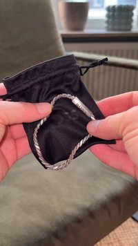 Ezüst színű, csavart lánc karkötőt tartó kezek, amelyen a "BOSS" felirat olvasható, egy kis fekete húzózsinóros tasak felett, puha felületen.